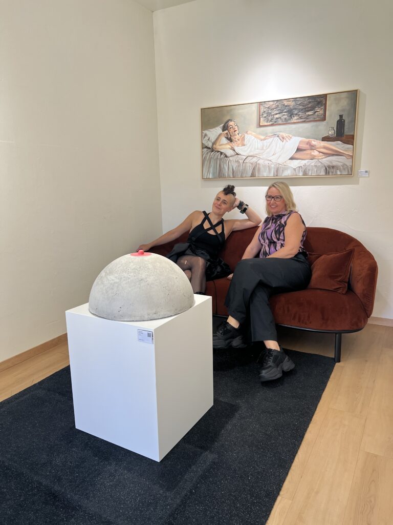 Two people on a sofa between a concrete sculpture and a painting.